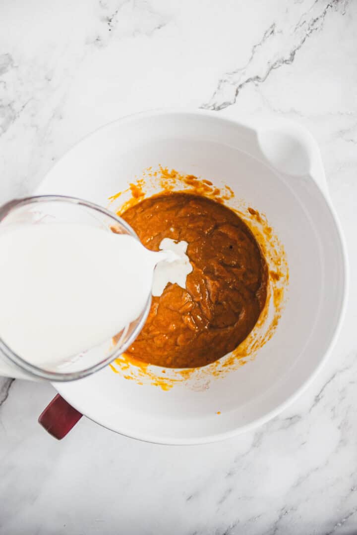 Milk being poured into a mixing bowl with orange batter on a white marble countertop.
