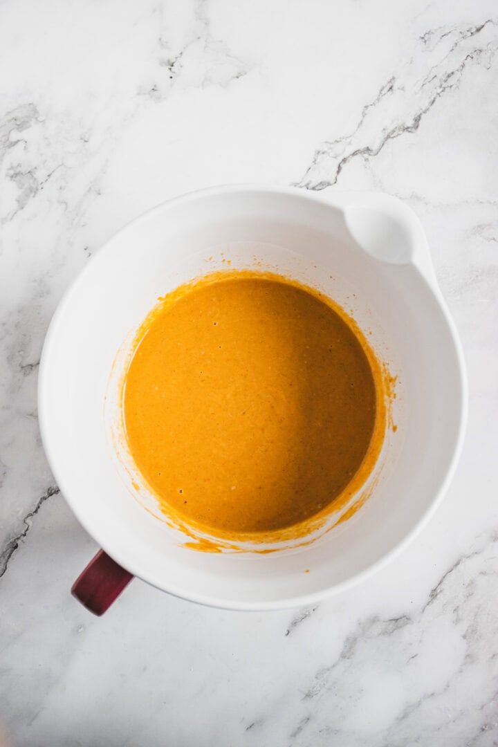 White mixing bowl with orange batter on a white marble surface, viewed from above.