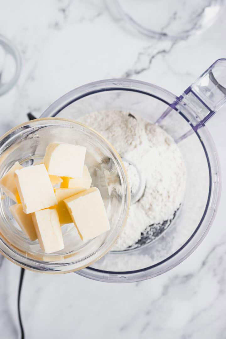 Cubes of butter in a glass bowl held over a food processor with flour inside on a marble countertop.