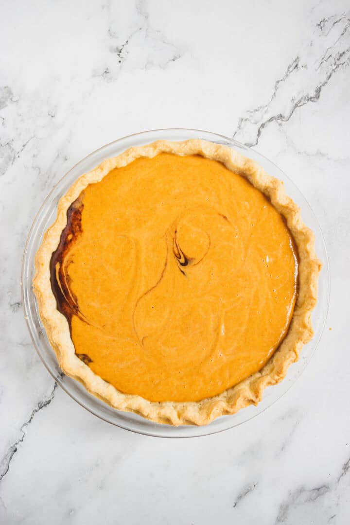 Pumpkin pie with a flaky crust in a glass pie dish on a white marble surface.