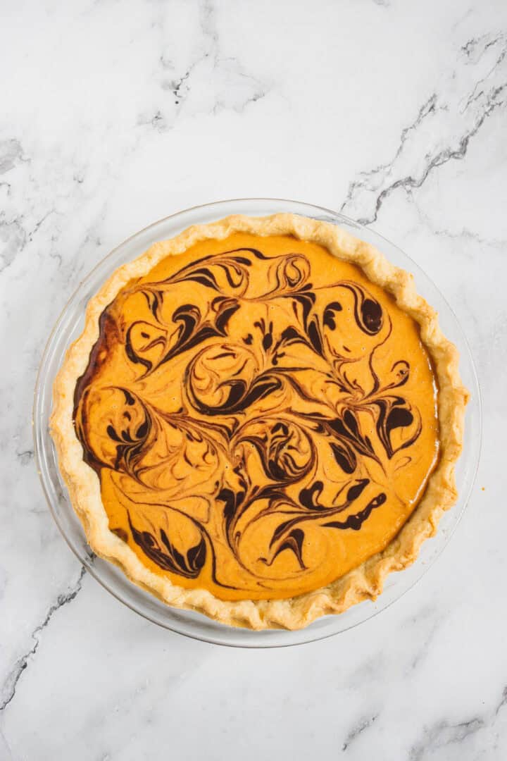 Pumpkin pie with a chocolate swirl design on top, in a glass pie dish on a marble surface.