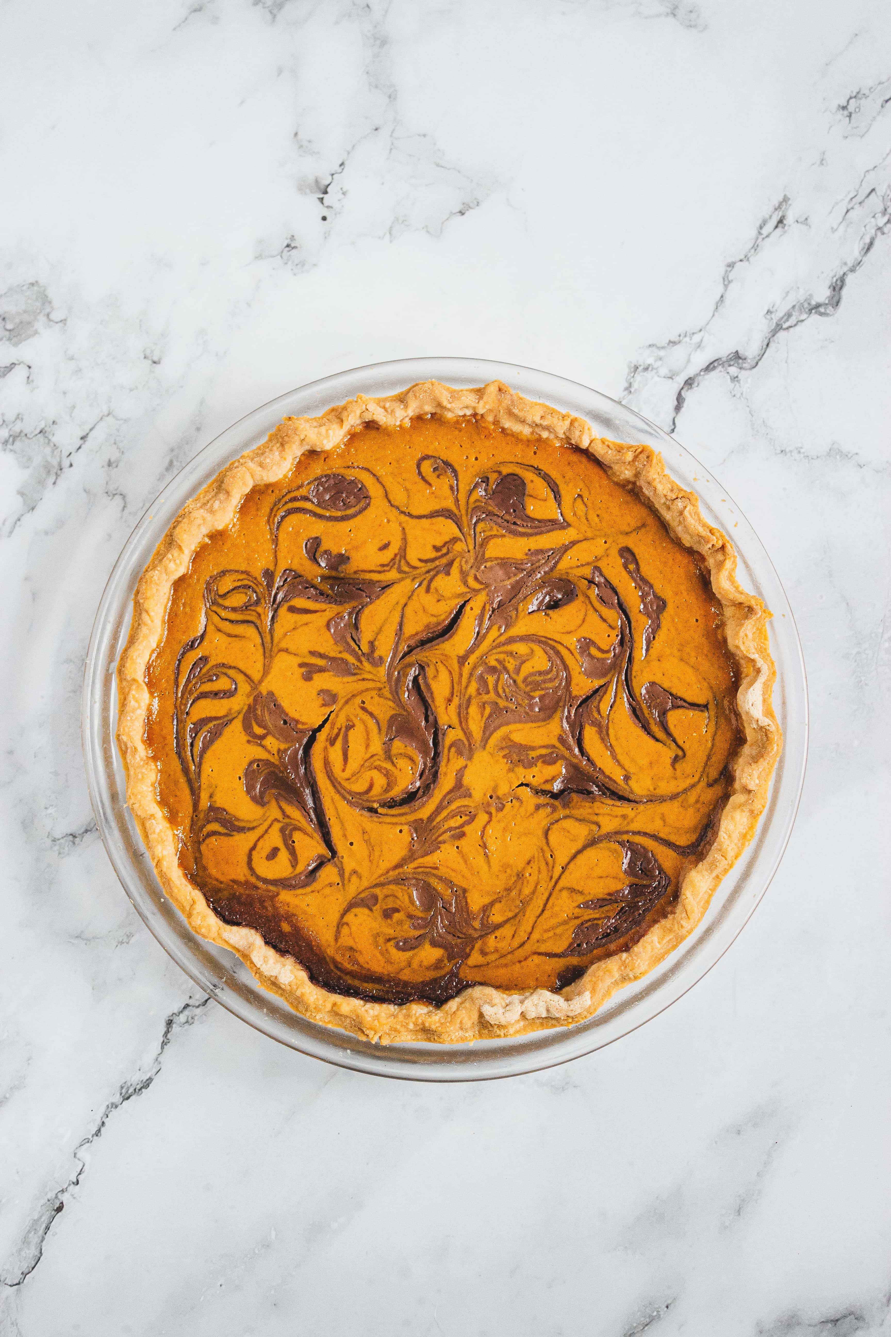 Pumpkin pie with a swirled chocolate pattern on top in a glass pie dish, set on a marble surface.