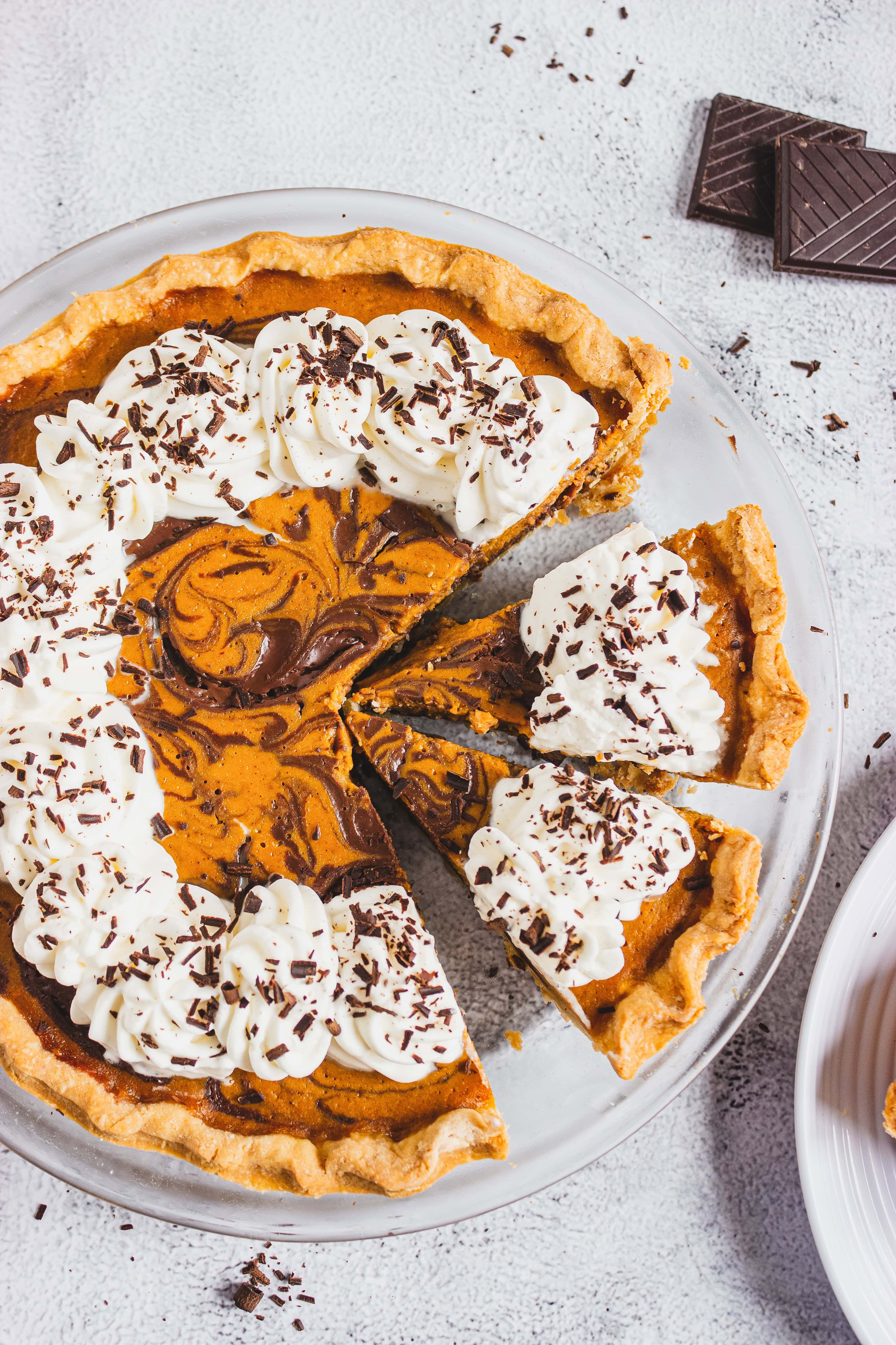 Pumpkin pie with chocolate swirls, whipped cream, and chocolate shavings on a white plate.