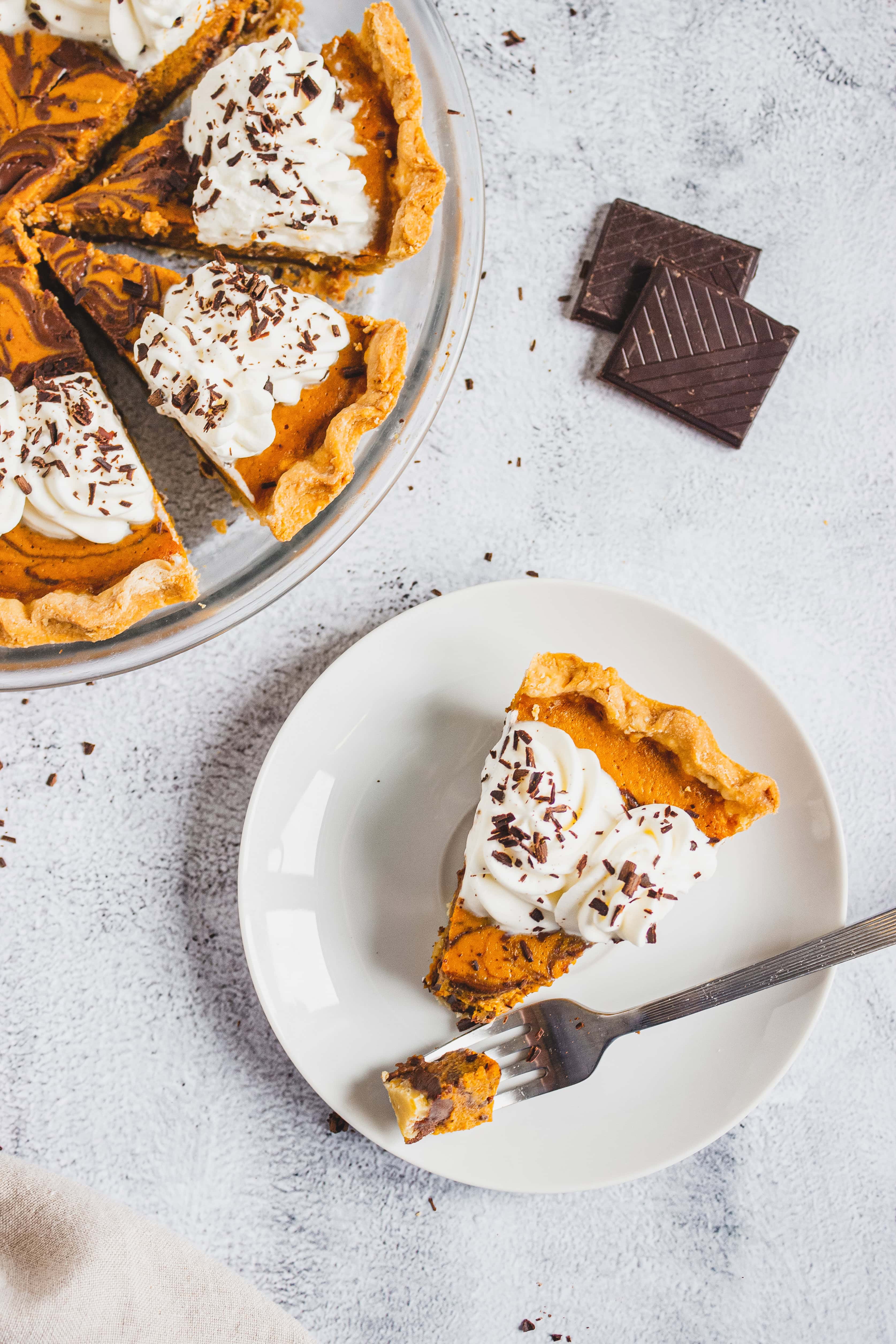 A slice of pumpkin pie with whipped cream and chocolate shavings on a plate beside the whole pie.
