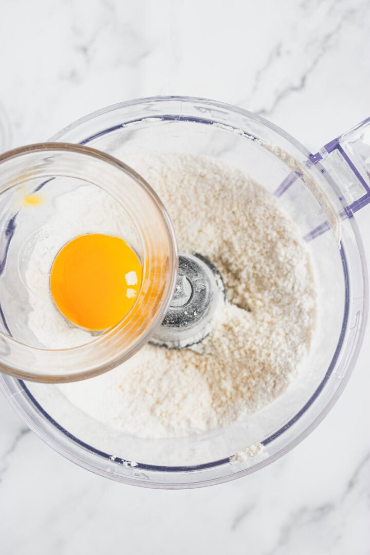 A bowl with an egg yolk held above flour in a food processor on a marble countertop.