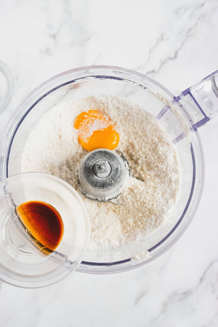 Top view of a food processor with flour and an egg yolk, as vanilla extract is being poured in.