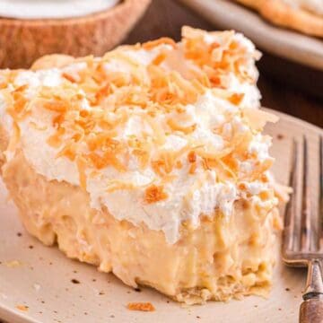 A slice of coconut cream pie topped with whipped cream and toasted coconut on a beige plate with a fork.