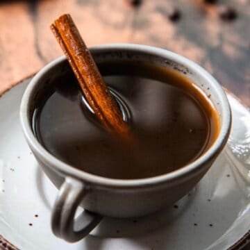 A cup of dark coffee with a cinnamon stick inside, placed on a white saucer.