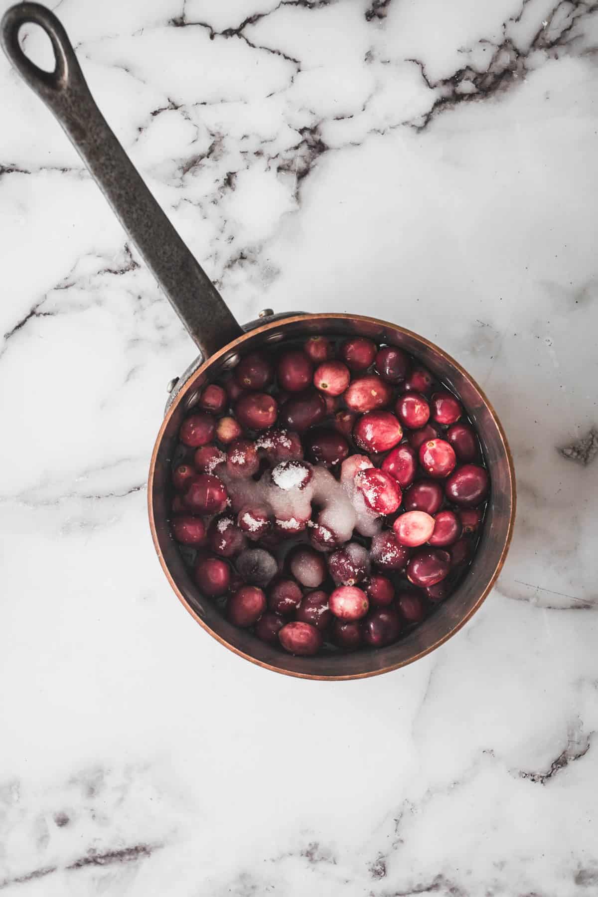 A saucepan filled with cranberries and sugar sits on a white marble surface, ready to be transformed into a delicious cranberry dip.