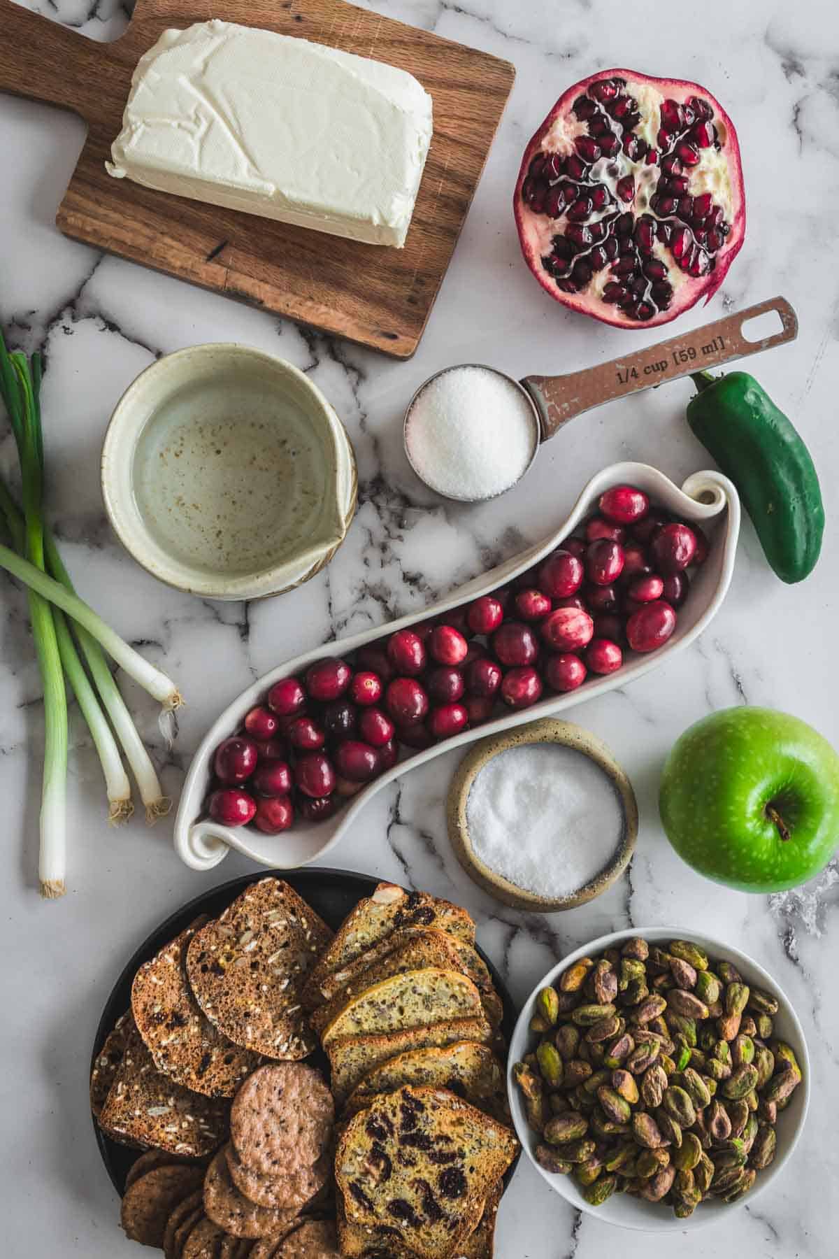 Ingredients on a marble surface: cream cheese, pomegranate, cranberries, green apple, crackers, pistachios, and seasonings.