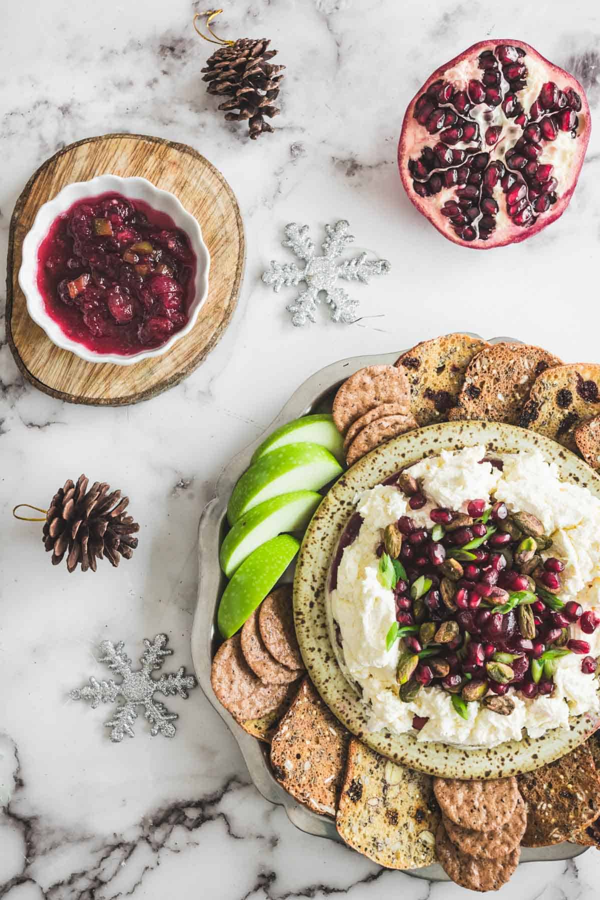 Festive cheese platter with crackers, green apple slices, pomegranate, and a tangy cranberry dip on a marble table.