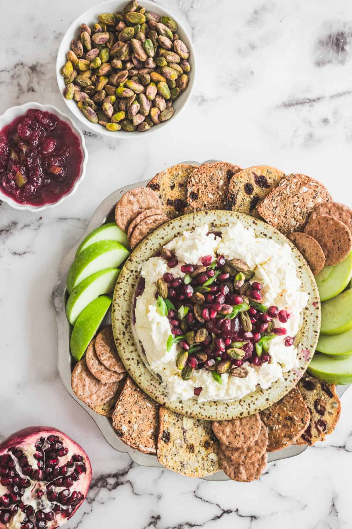 A platter with crackers, green apple slices, and cheese dip topped with pomegranate seeds and pistachios.