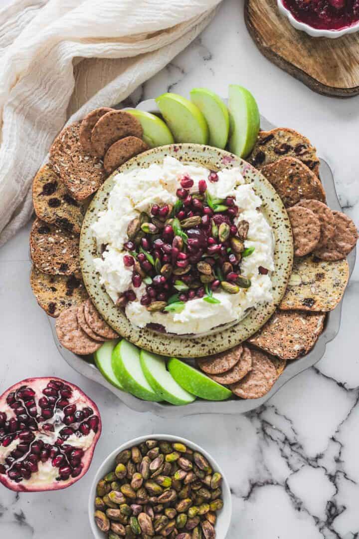 A plate with cheese topped with pomegranate seeds and pistachios, surrounded by crackers and green apple slices, is perfect served alongside a tangy cranberry dip.