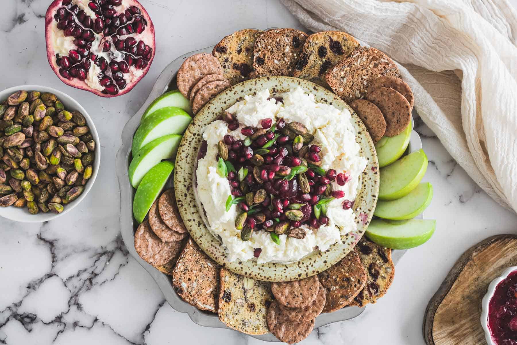 A plate of crackers, green apple slices, and cheese dip topped with pomegranate seeds and pistachios.
