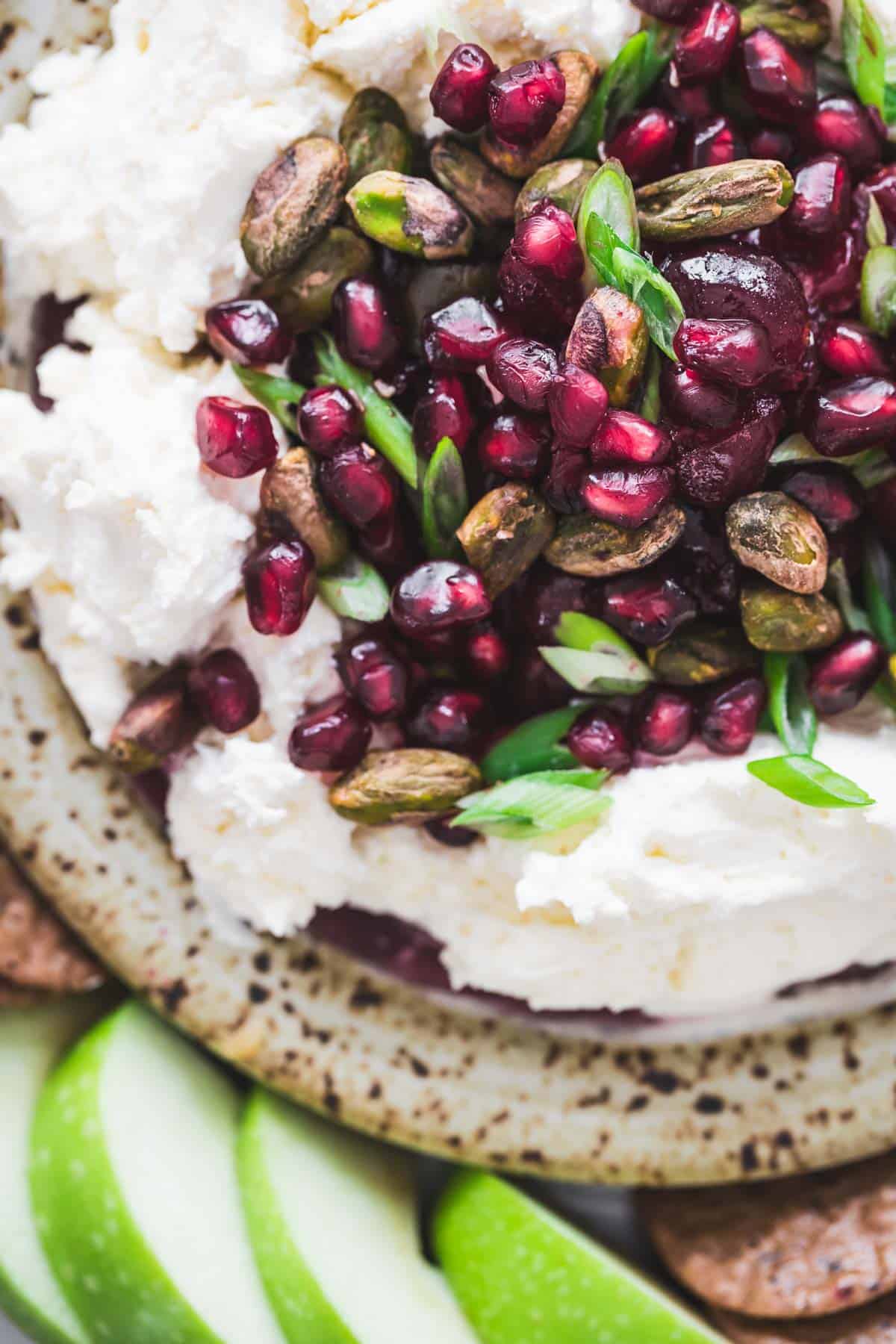 Close-up of whipped cheese topped with pomegranate seeds, pistachios, and green onions, with apple slices nearby.