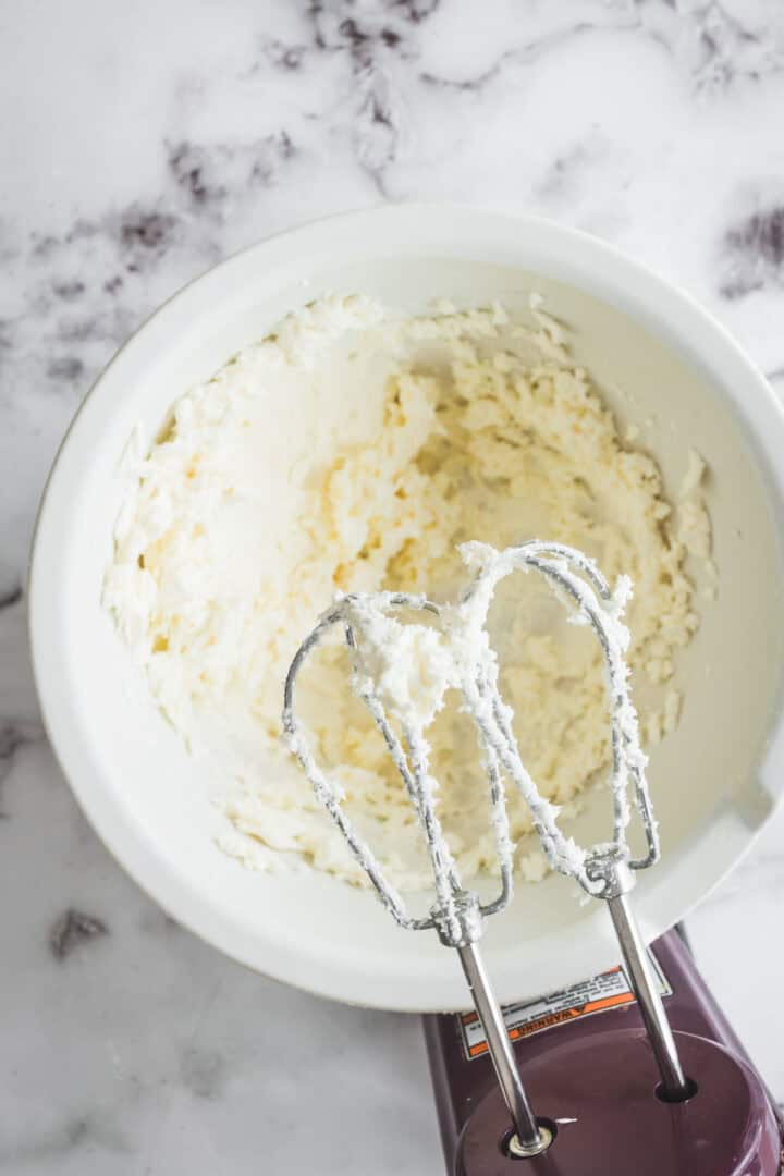 Electric hand mixer with whipped cream cheese in a white bowl on a marble countertop, perfect for preparing cranberry dip.