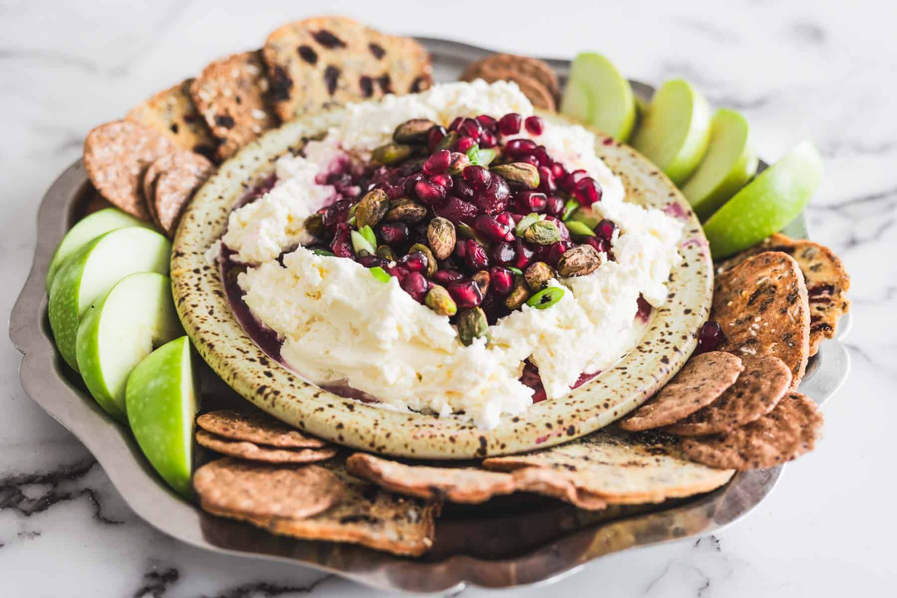 A plate of ricotta cheese topped with pomegranate seeds and pistachios, surrounded by crackers and green apple slices.
