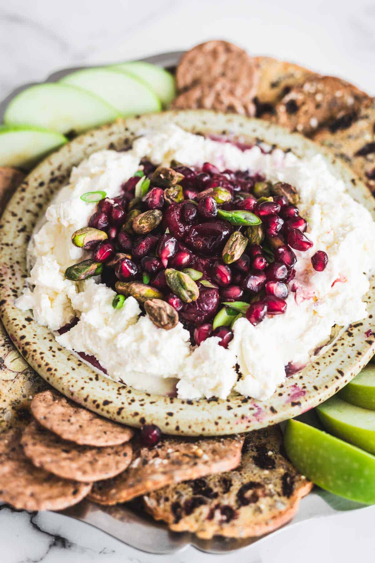 A plate of whipped ricotta, reminiscent of a cranberry dip, topped with pomegranate seeds and pistachios, surrounded by crackers and apple slices.