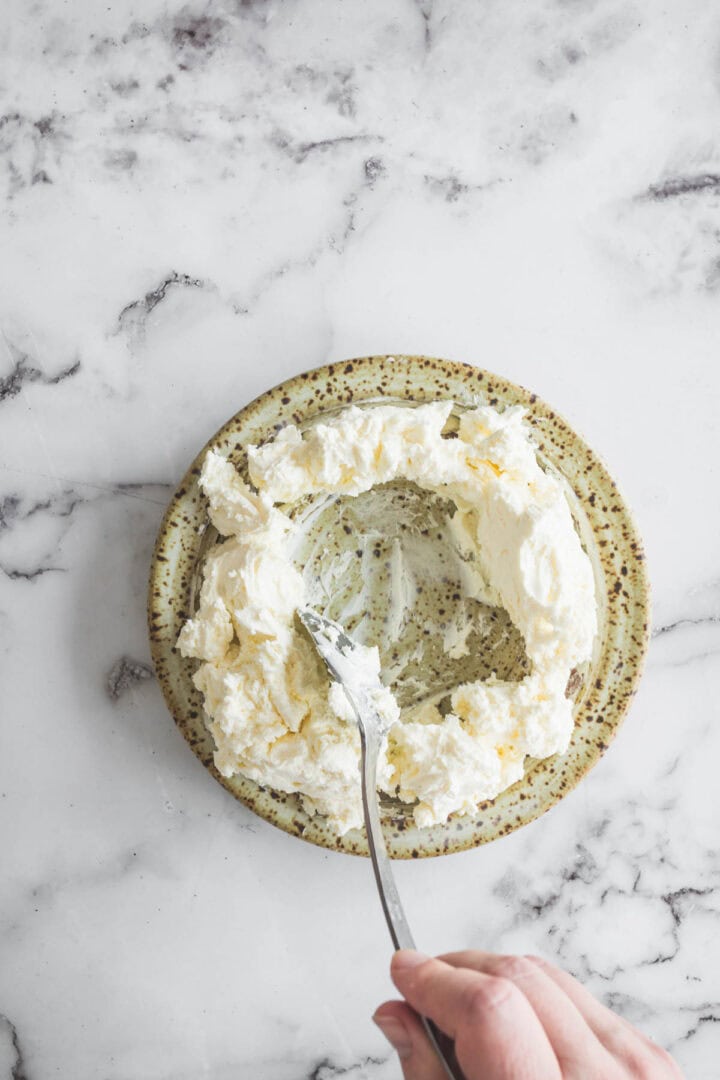 A hand holding a fork mashes creamy cheese for cranberry dip on a speckled plate atop a marble surface.