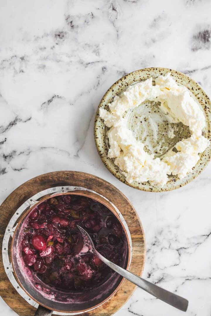 Overhead view of whipped cream in a bowl and a pot of cherry compote with a spoon on a marble surface, perfect for pairing with your favorite cranberry dip.