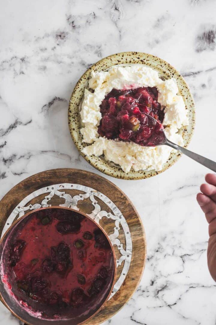 A hand spoons berry compote onto whipped ricotta on a plate, with more cranberry dip in a bowl nearby.