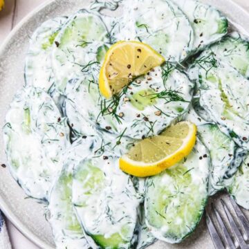 Sliced cucumber salad with creamy dressing, fresh dill, and lemon wedges on a white plate with a fork.