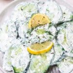 Sliced cucumbers in creamy dill sauce, topped with black pepper and lemon wedges on a plate.