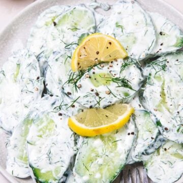 Sliced cucumbers in creamy dill sauce, topped with black pepper and lemon wedges on a plate.
