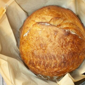 Round golden loaf of bread on parchment paper in a black cast iron pot, viewed from above.