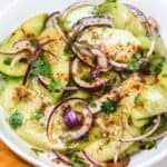 Mexican Cucumber Salad features sliced cucumbers, red onions, and cilantro topped with chili powder and seasonings in a white bowl.