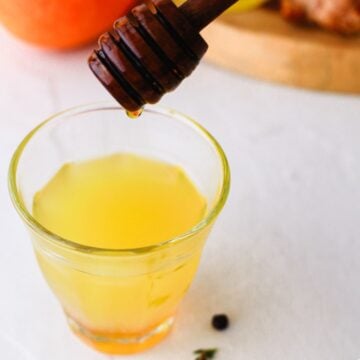 A honey dipper drips honey into a glass of yellow liquid on a white surface.