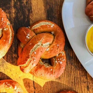 Soft pretzels with coarse salt on a wooden surface, with mustard dip on a white plate nearby.