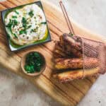 Fried potato sticks in a basket, creamy dip in a bowl, and chopped herbs on a wooden board.