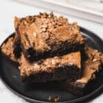 Three chocolate brownies with a nut butter swirl and seeds stacked on a black plate.