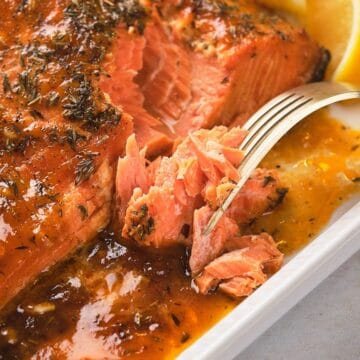 Close-up of glazed baked salmon being flaked with a fork, served with lemon wedges on a white plate.