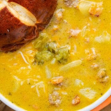 A bowl of cheesy soup with ground meat, potatoes, and vegetables, served with a pretzel bun.
