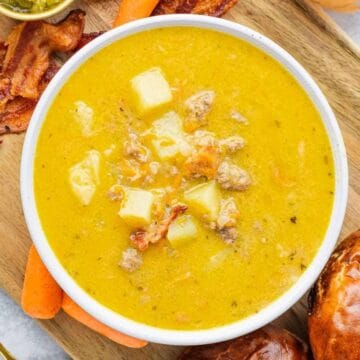 A bowl of cheesy soup with potatoes, served with bacon, carrots, and bread on a wooden board.