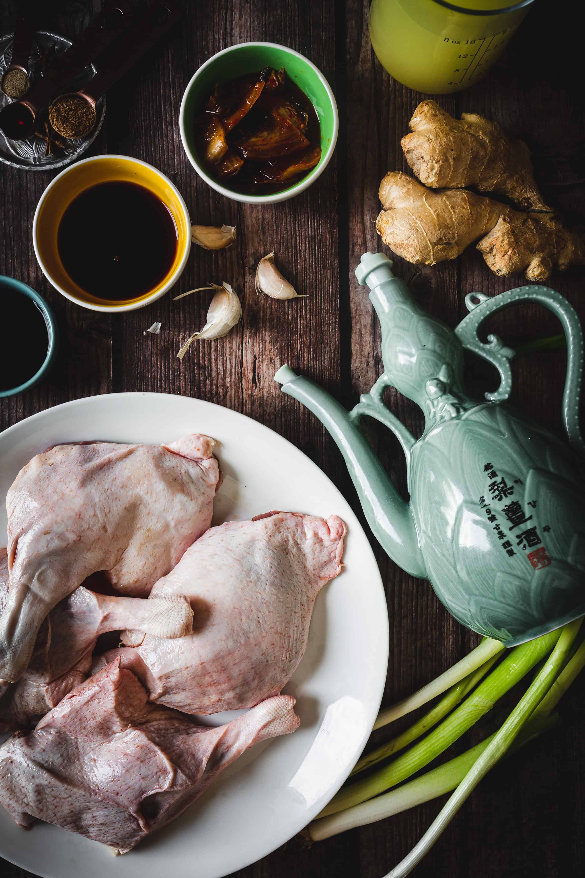 Raw duck pieces on a plate, surrounded by ginger, garlic, green onions, sauces, and a decorative teapot.