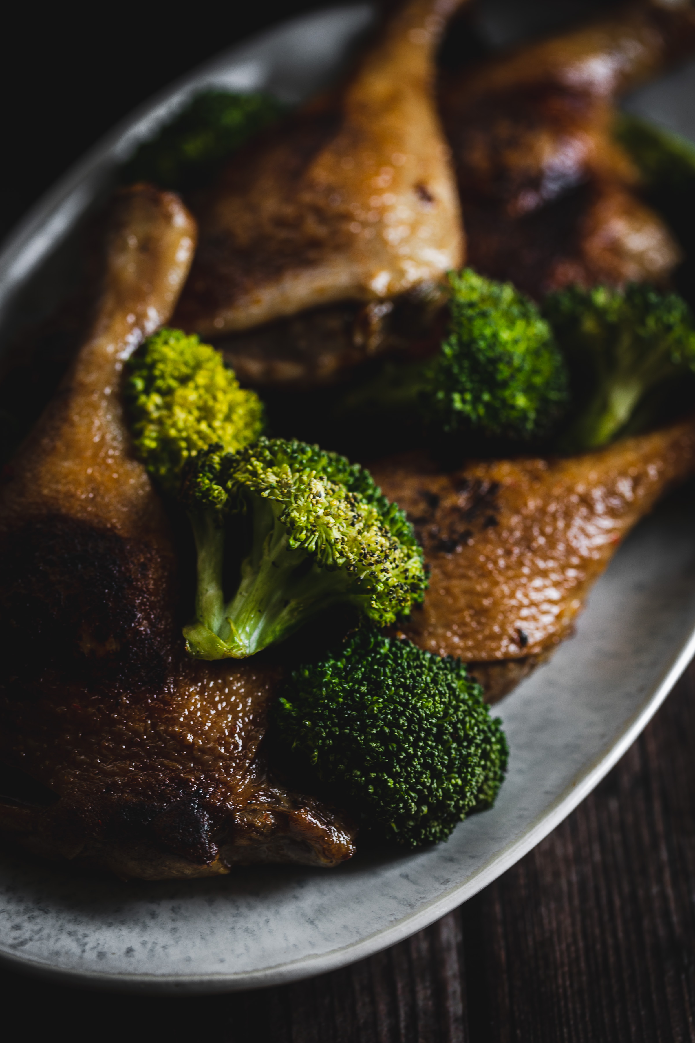 Roasted duck legs with broccoli florets on a white plate, served on a dark wooden table.