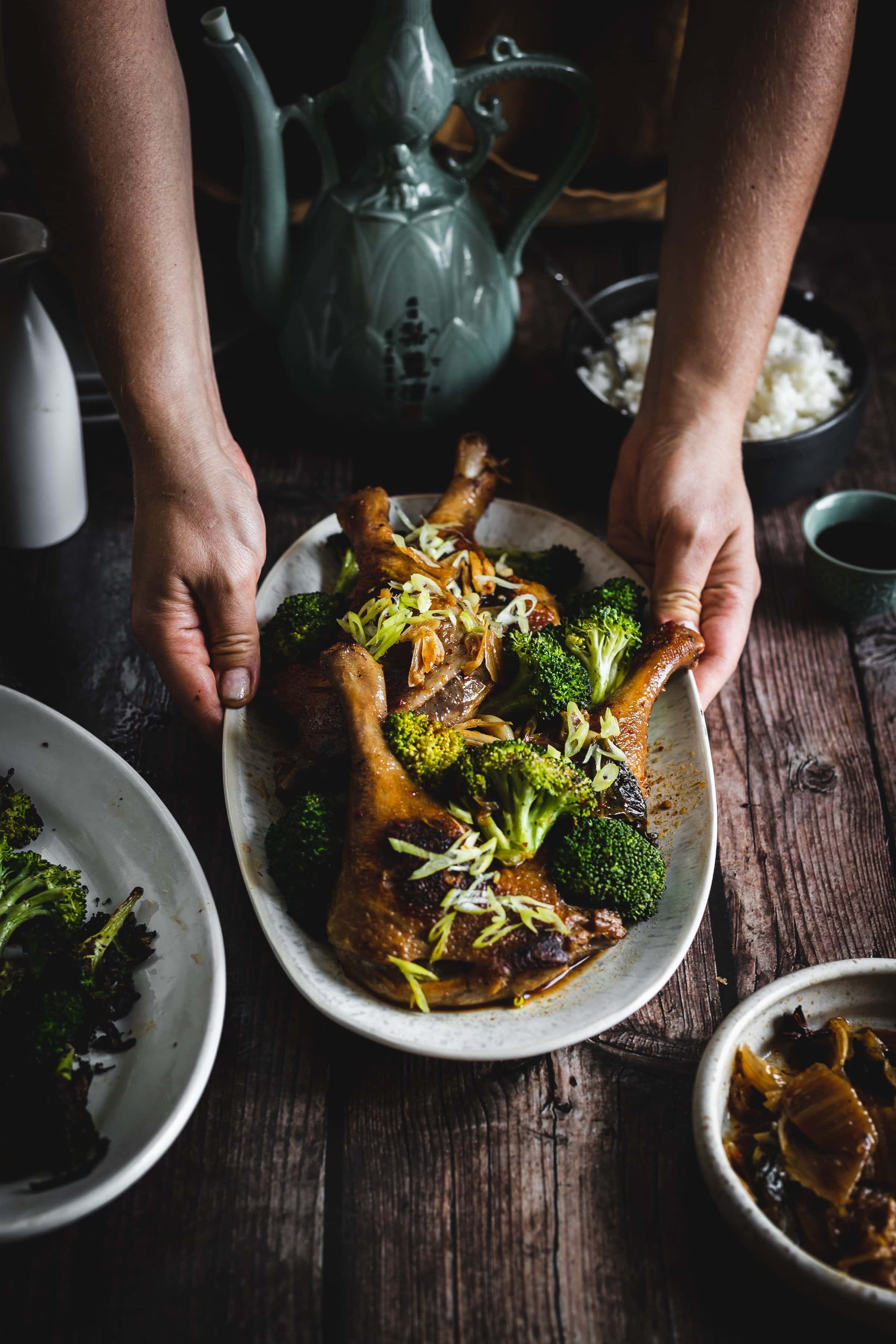 Hands holding a platter of roasted chicken legs with broccoli, garnished with sliced green onions and garlic.