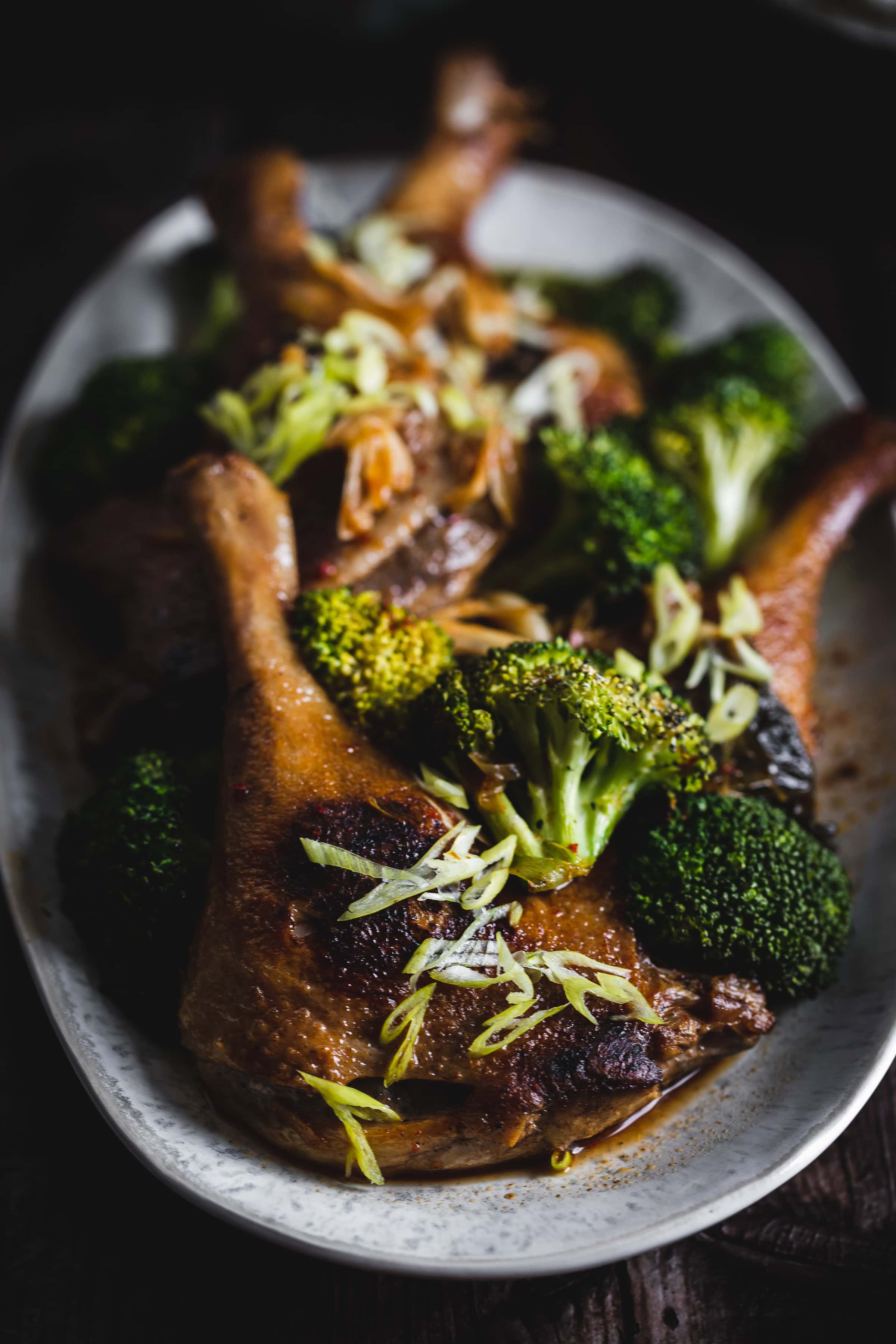 Roast duck legs garnished with sliced green onions, served with steamed broccoli on a white oval plate.