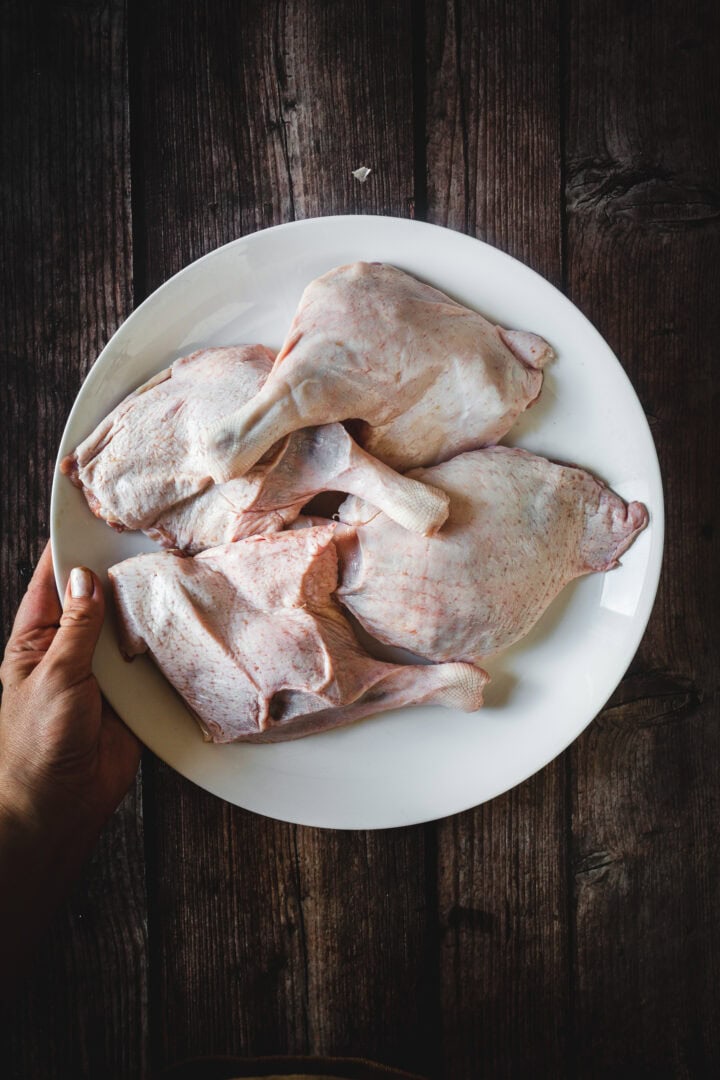 A hand holds a white plate with four raw duck legs on a dark wooden surface.