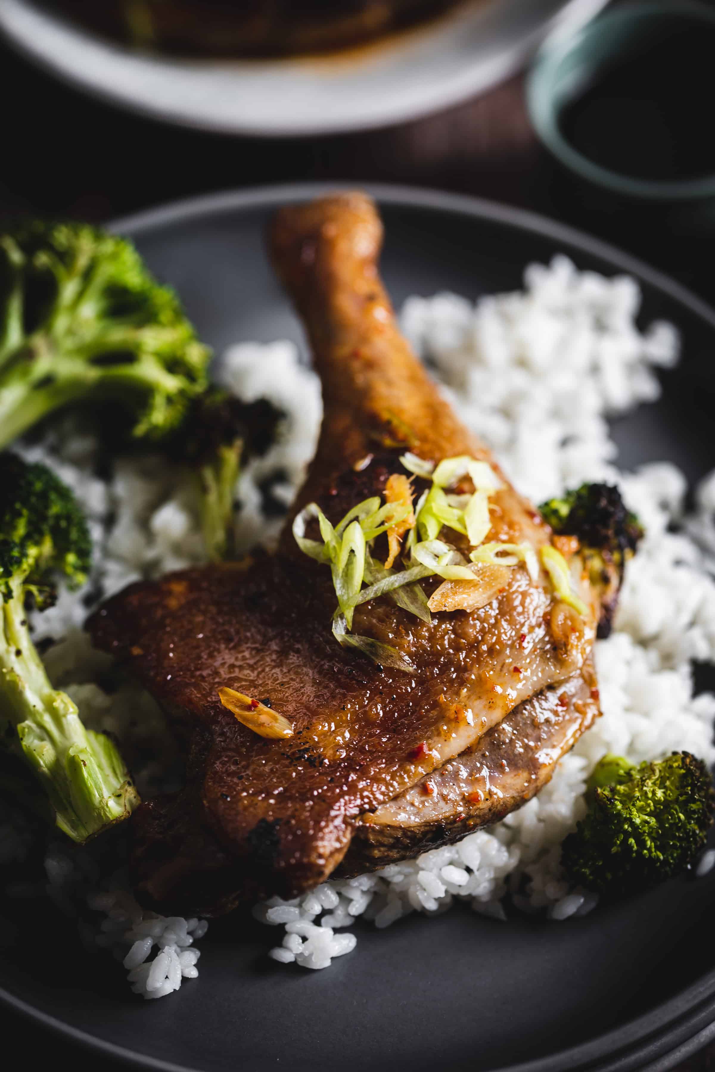 A plate of rice topped with roasted duck leg, garnished with green onions, and served with broccoli.