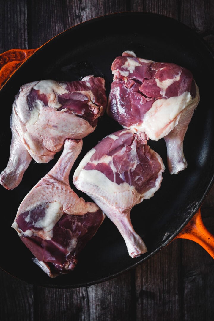 Four raw duck legs arranged in a cast iron pan on a dark wooden surface.