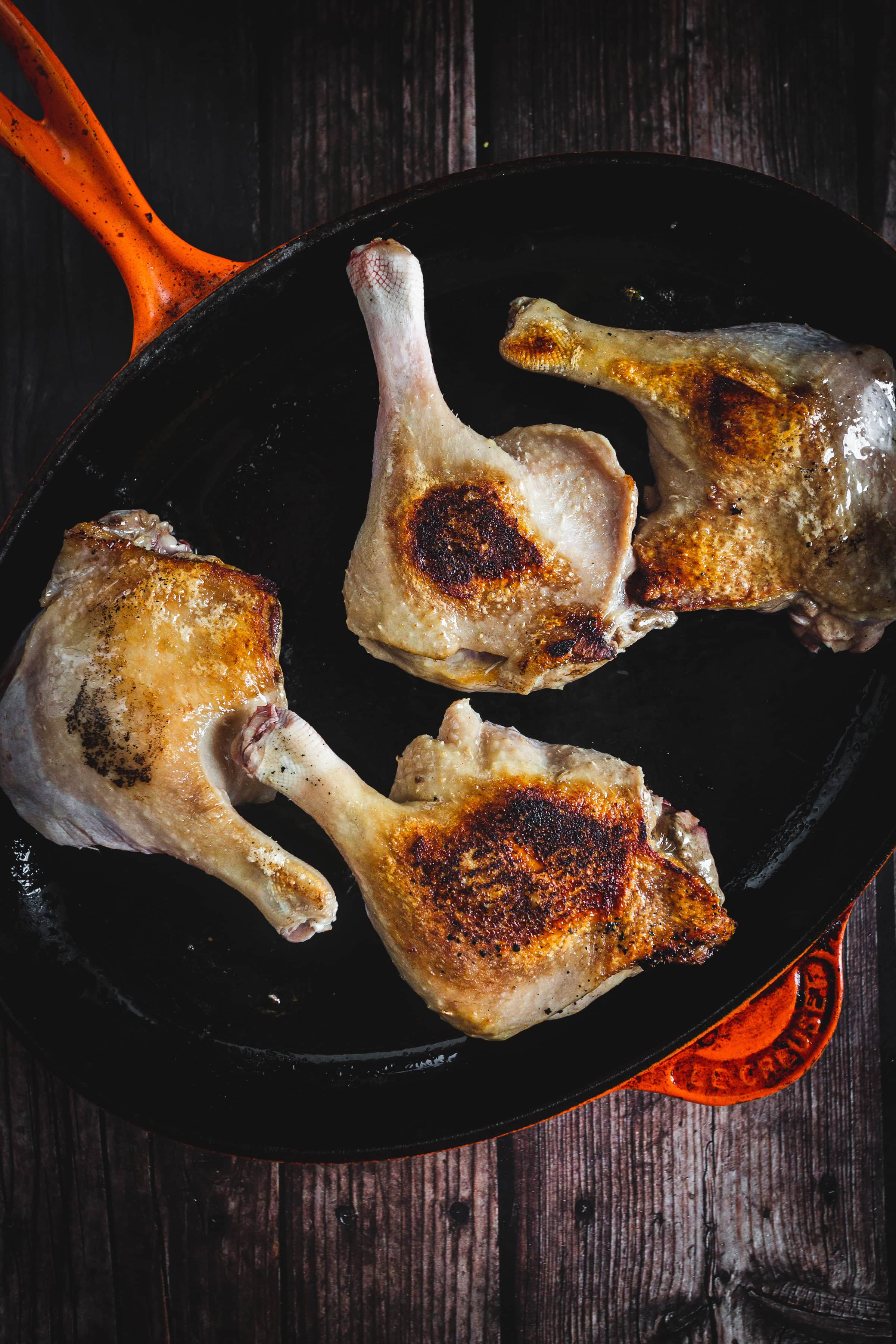 Four browned duck legs cooking in a black cast iron skillet with an orange handle on a dark wooden surface.
