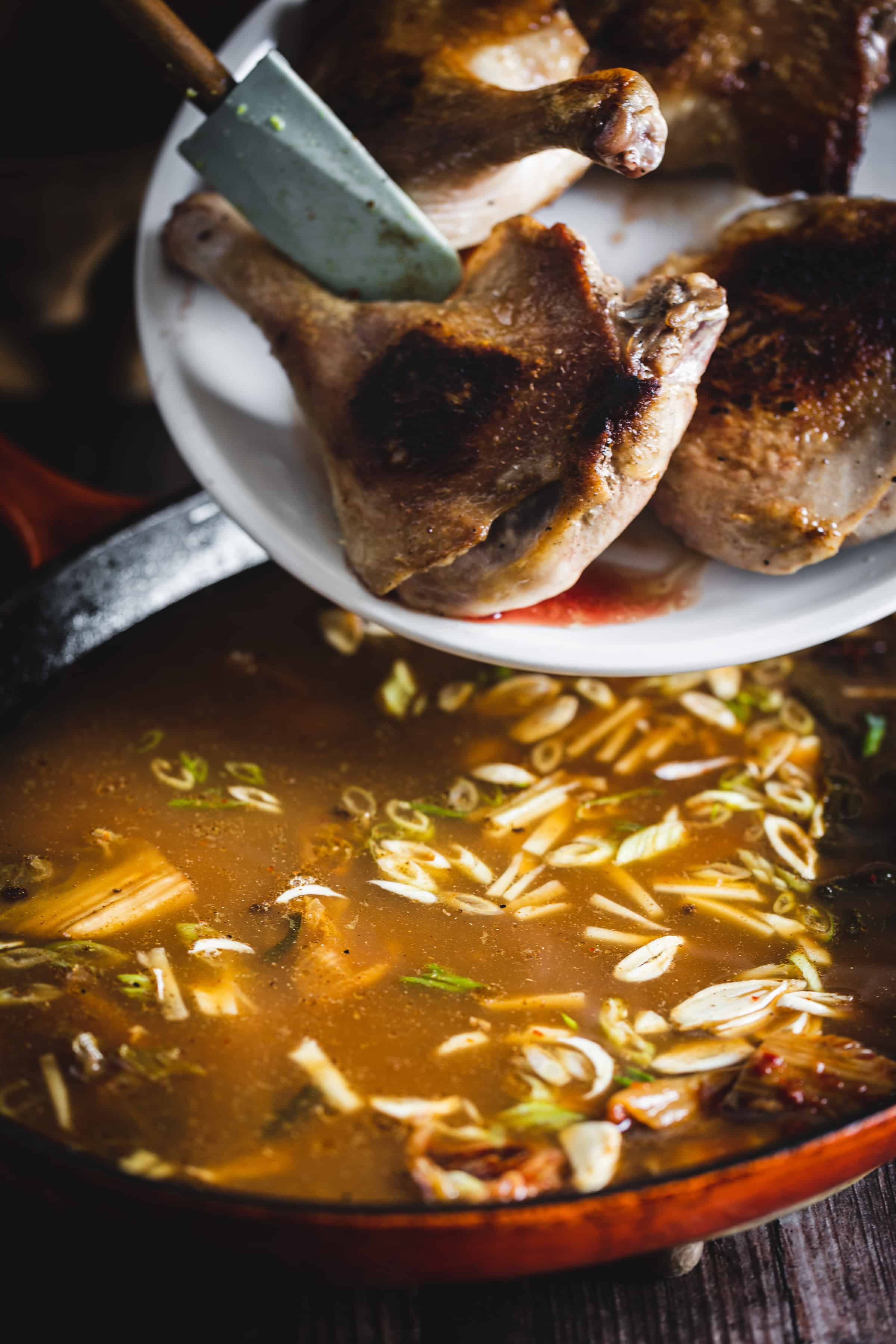 A plate of cooked duck legs is being added to a pot of broth with sliced green onions.