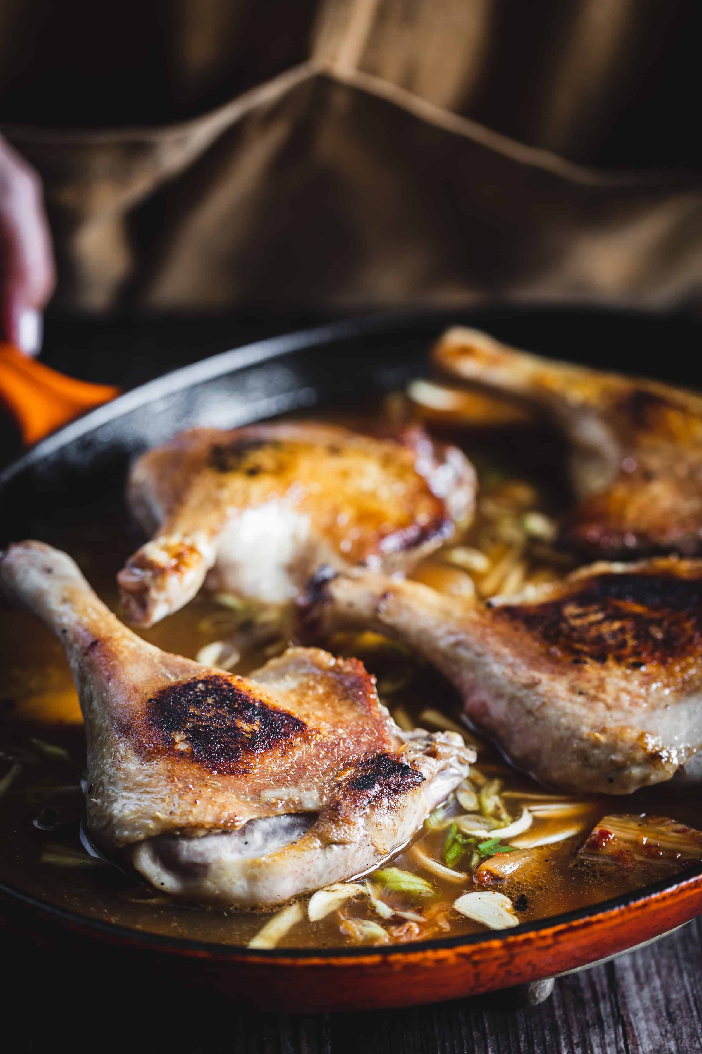 Four browned duck legs simmering in broth with sliced vegetables in a skillet.