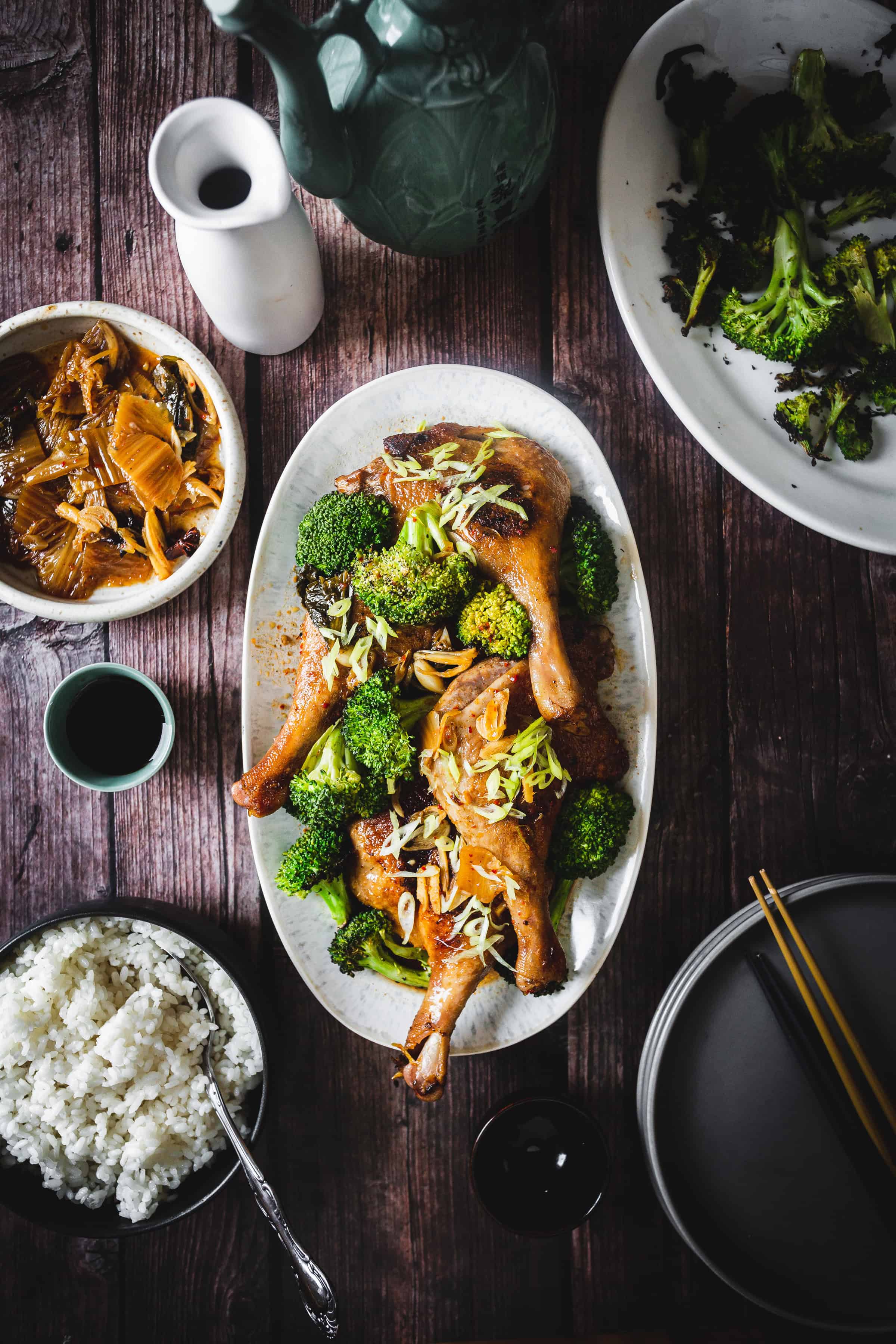 Oval plate of roasted chicken legs with broccoli, surrounded by rice, vegetables, and sauces on a wooden table.