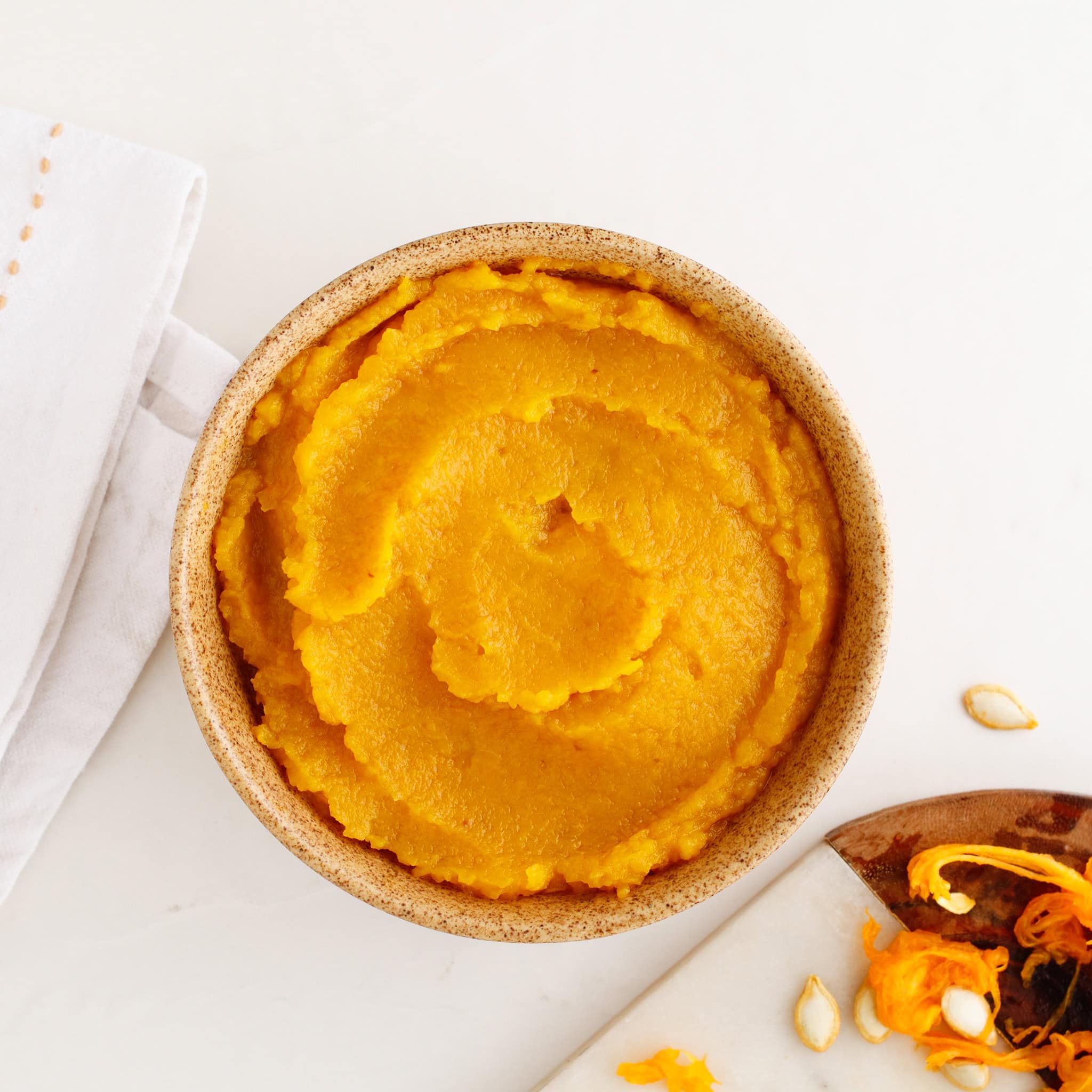 A bowl filled with smooth, vibrant pumpkin puree sits on a white surface next to a spoon and napkin.