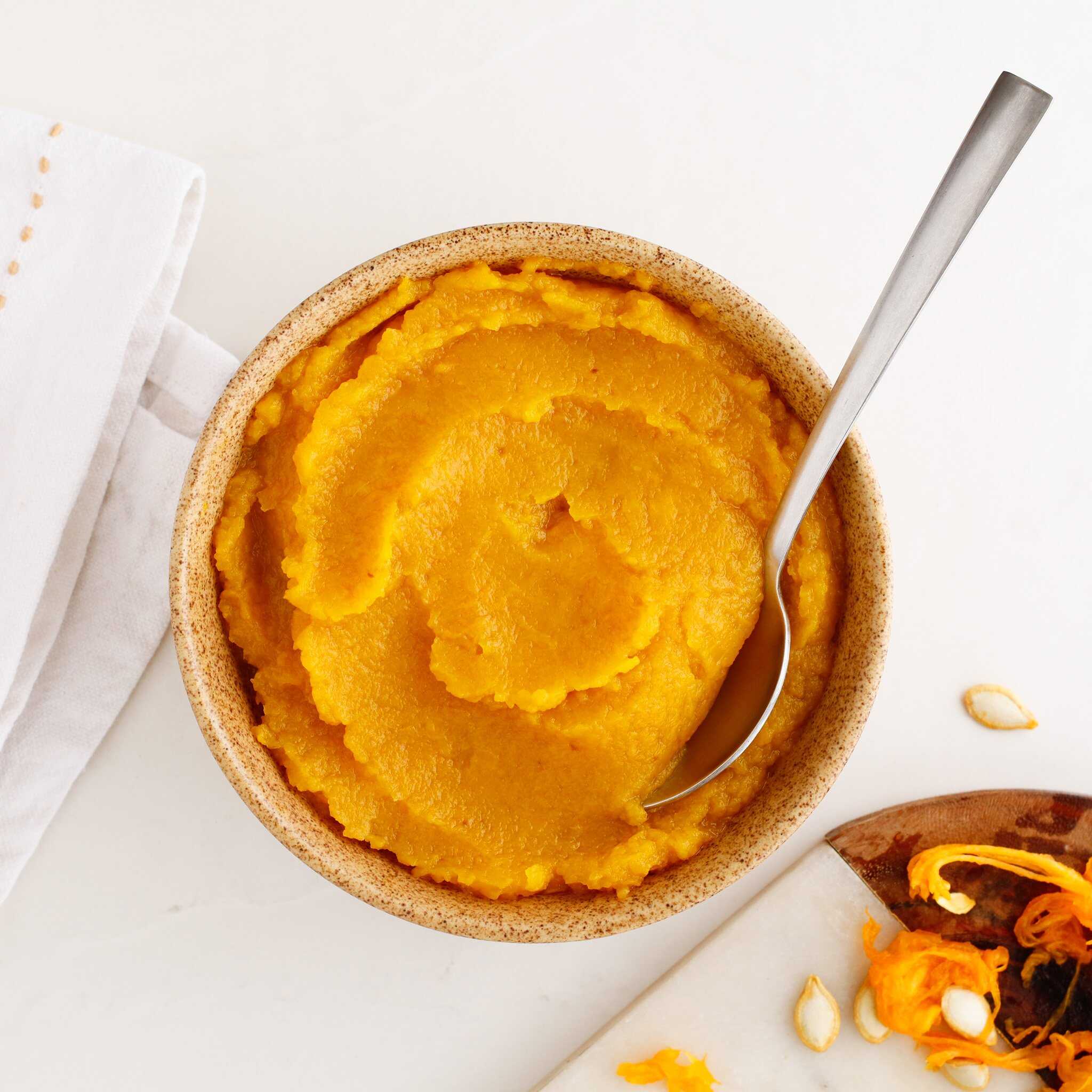 A bowl of creamy pumpkin puree with a spoon sits on a white surface, accompanied by a napkin and scattered pumpkin scraps.